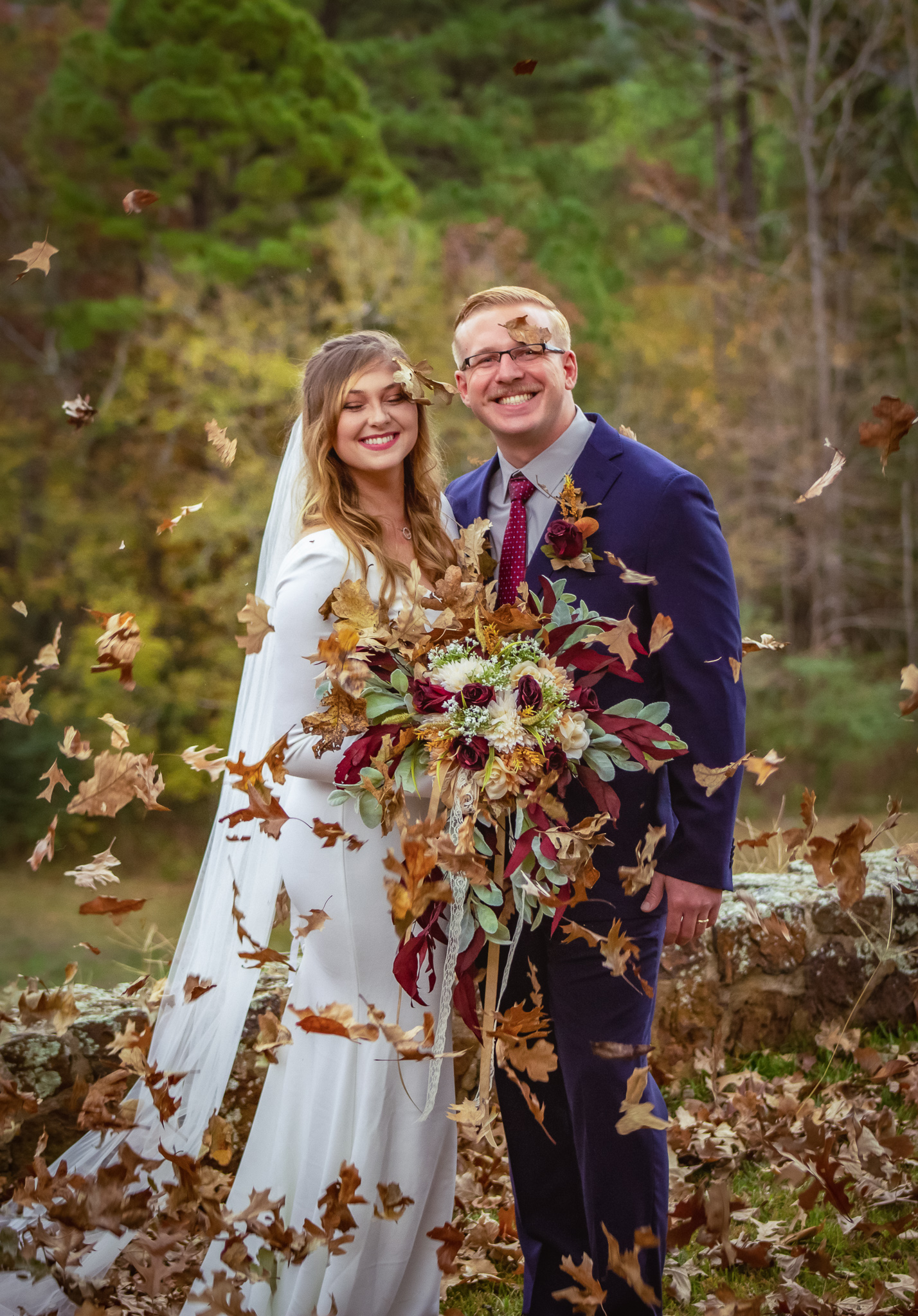 a bride and groom stand side by side smiling at the camera as a shower of crispy fall leaves fall from the sky