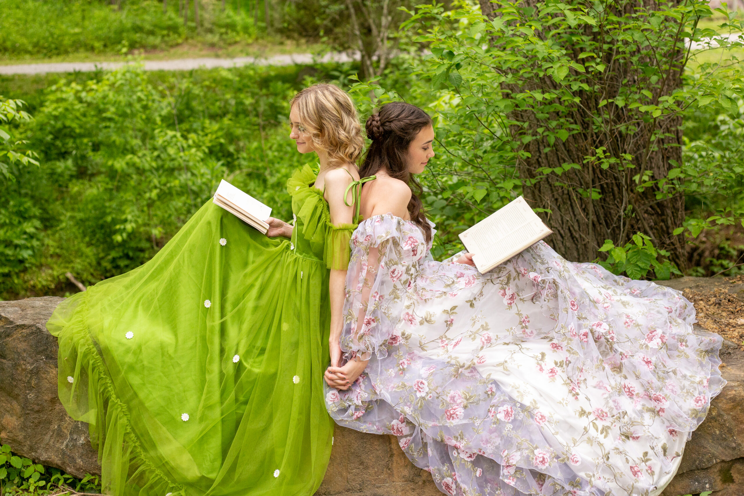 two sisters in princess dresses sit back to back on a stone wall each with knees propped to read a book in their lap, they hold hands as their dresses flow to the ground below them.