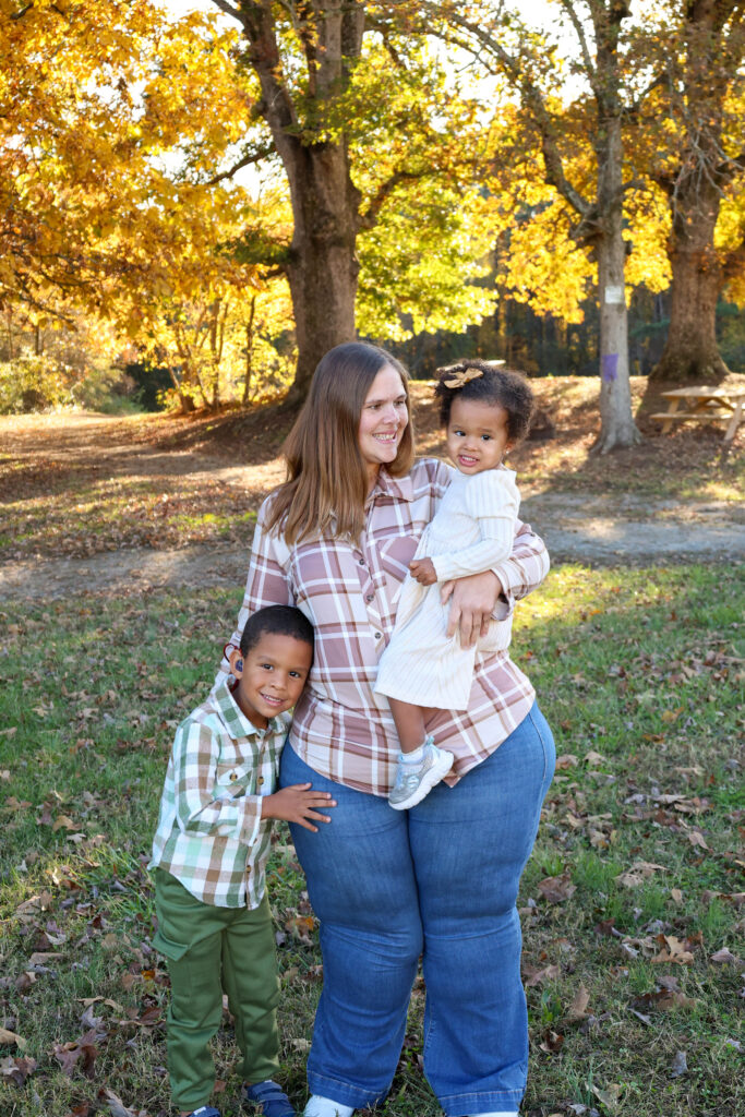 A mom and her two young children, boy and girl, standing in a small natural area with gorgeous orange trees in the background, the mom is smiling at her little girl in her arms, and her little boy in standing next to  her hugging her leg.