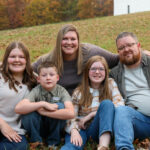 A family of 5 crouching and scooted next together in a field with a white barn in the background. All members are smiling and happy to capture this memory!