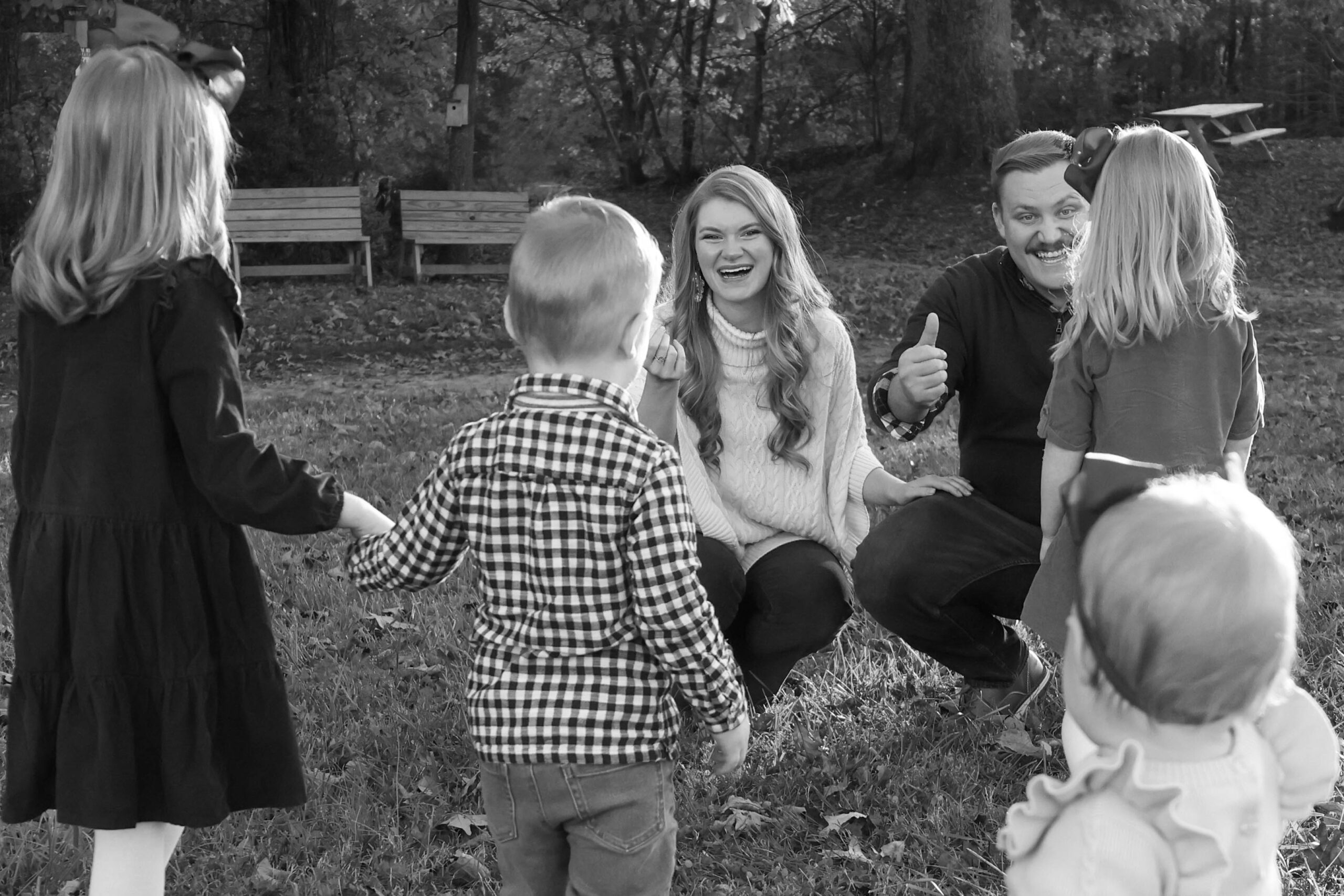 Family of six in a sunny field, with parents crouched down and their three daughters and young son running to them, creating a warm, heartfelt family scene.