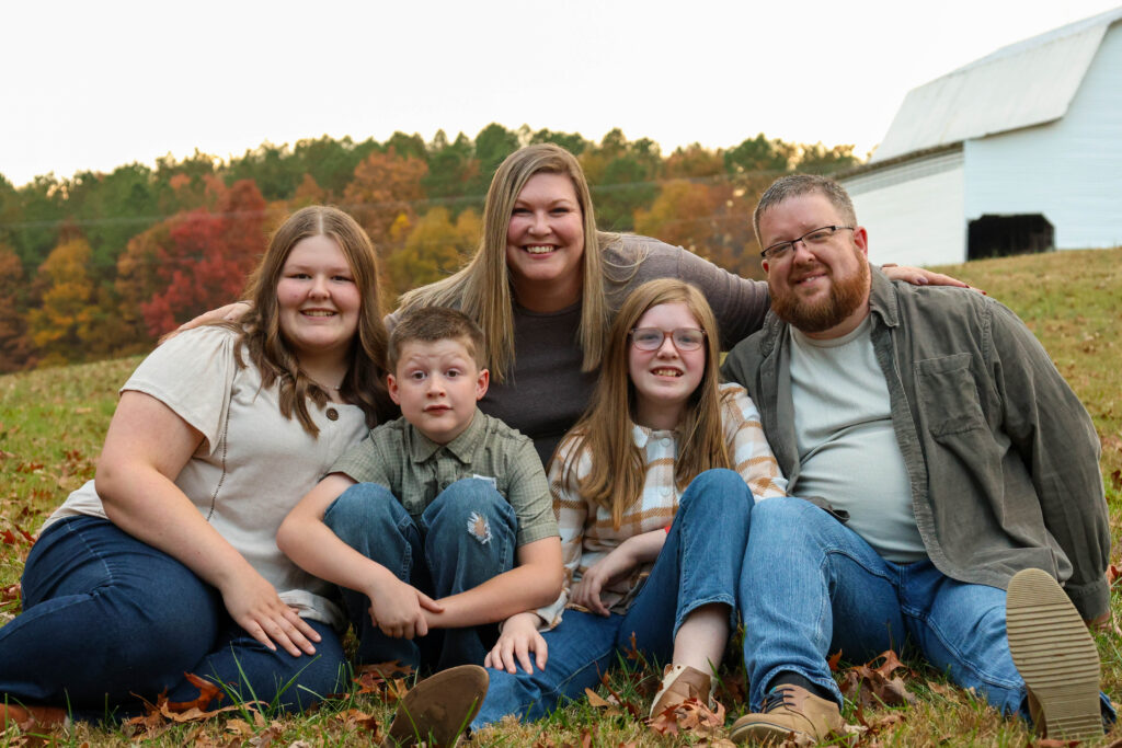 A family of 5 sit in a field gathered together and smiling