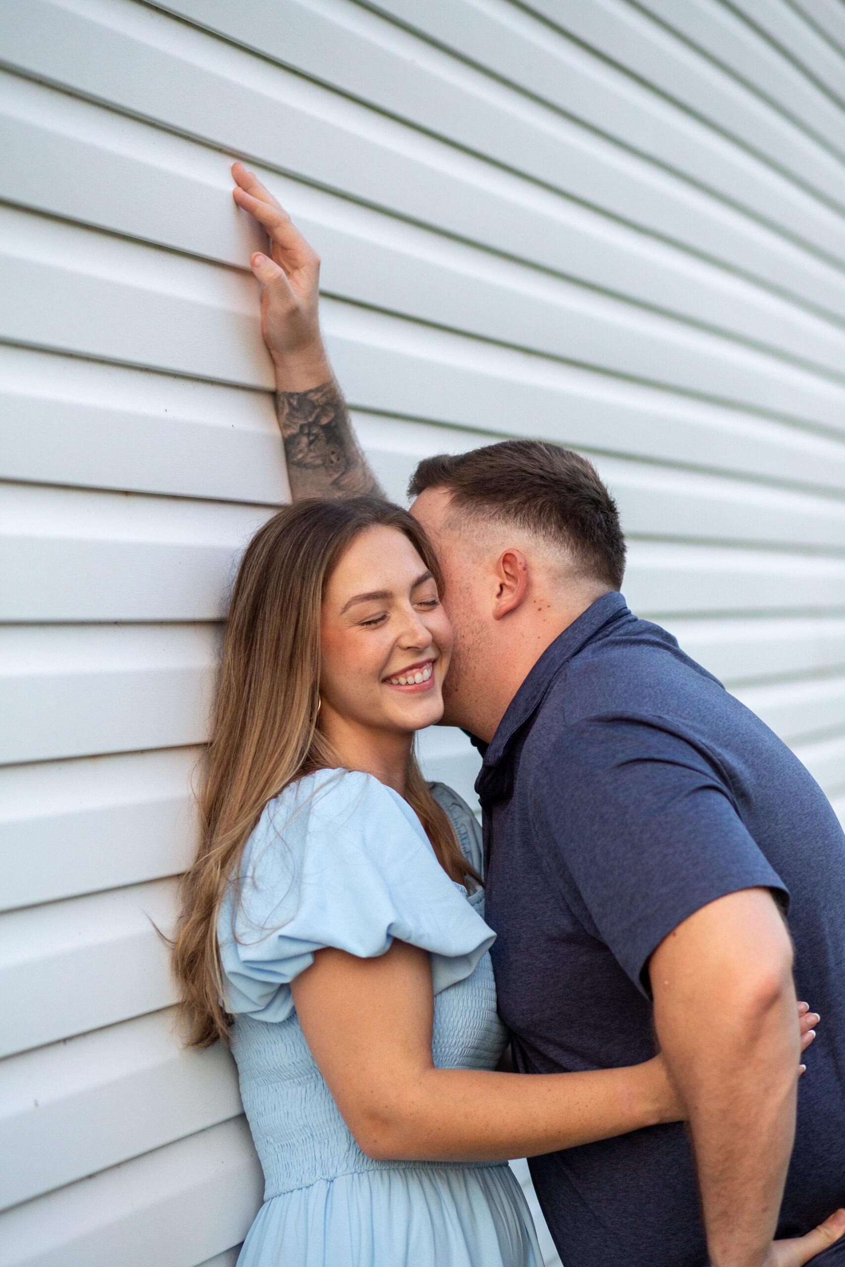 A husband leans against a white barn exterior, and whispers something funny into his wife's ear, she giggles and holds his waist as she enjoys his company.