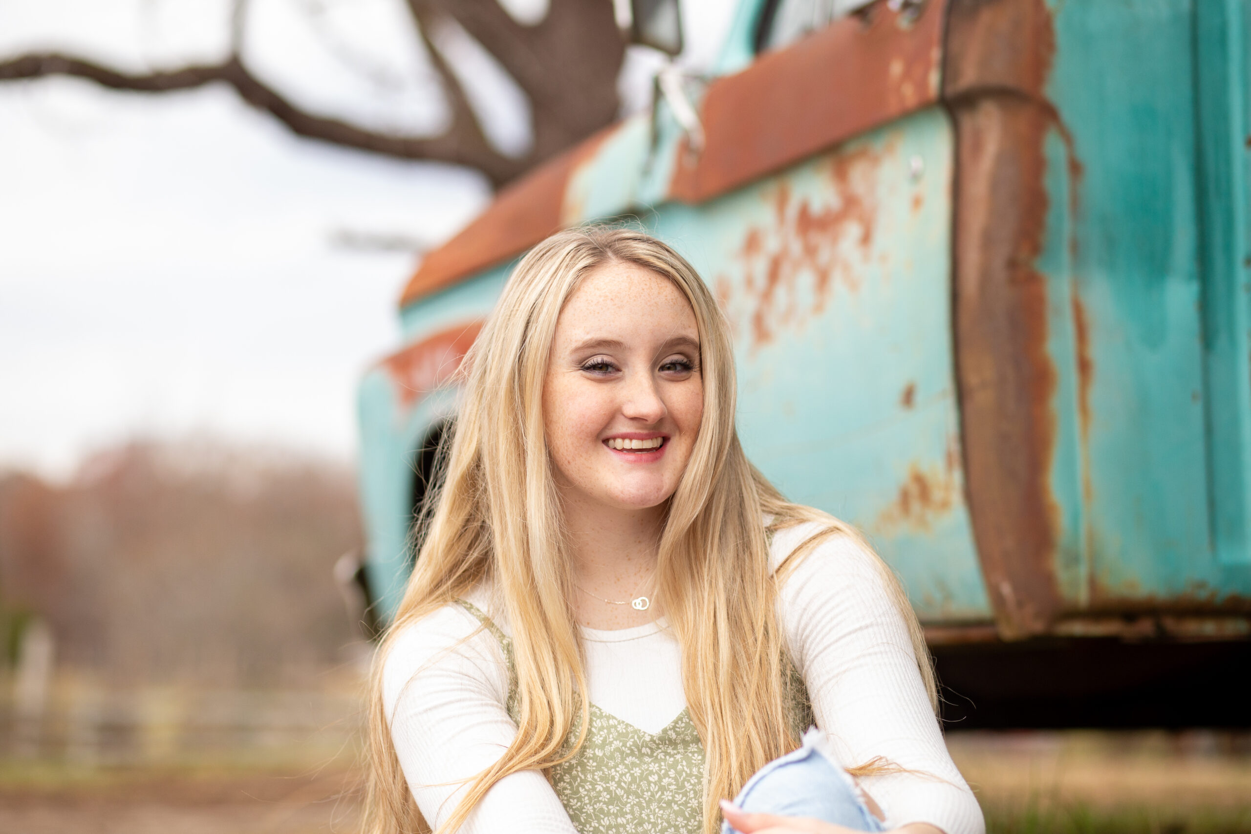 Denver NC Senior Photographer A Blonde haired and blue eyed senior sitting on the ground, in front of an old rusty pick up truck, smiling at the camera