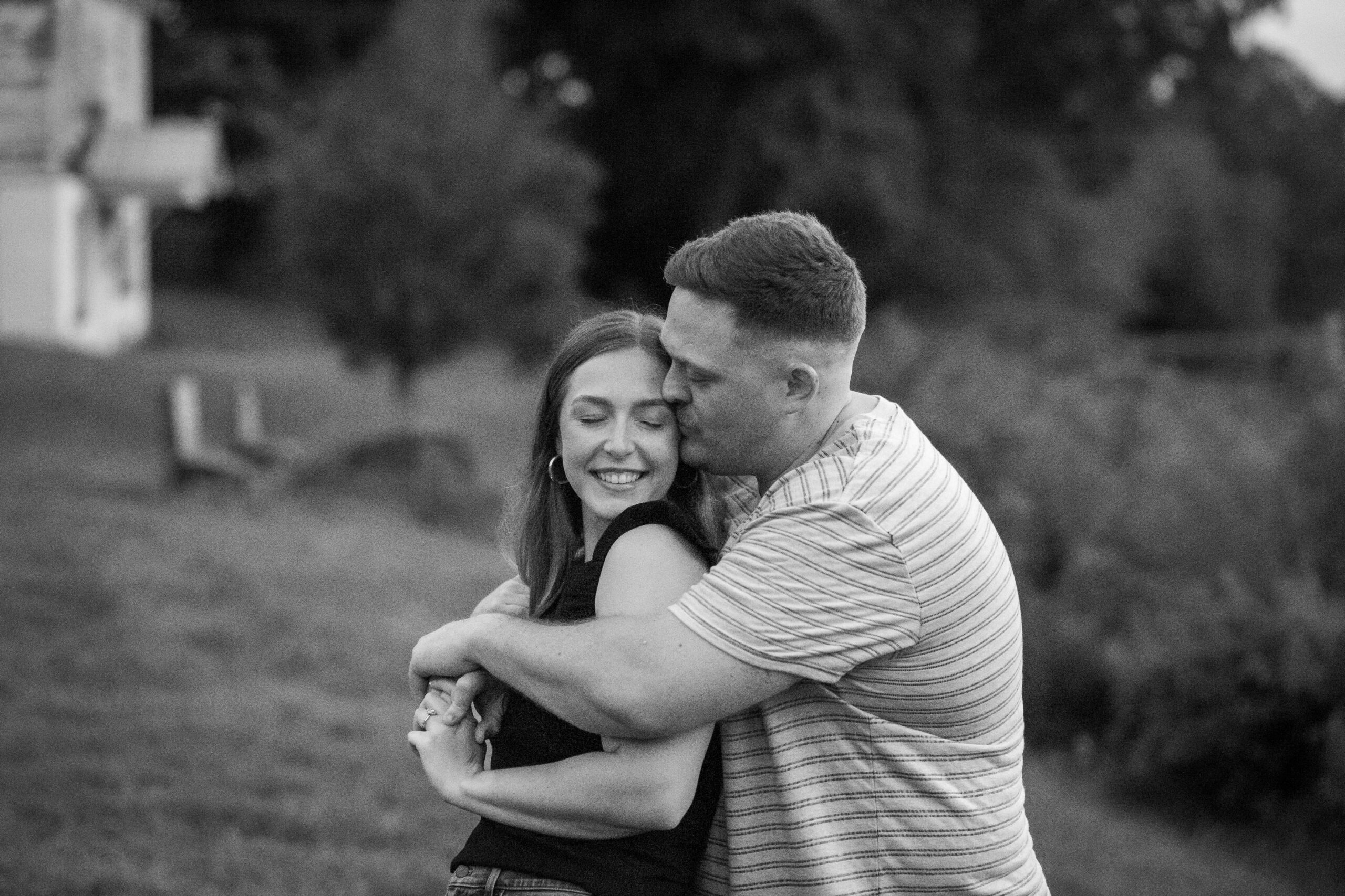 A husband embraces his wife from behind, holding her close and kissing her temple, She closes her eyes and smiles